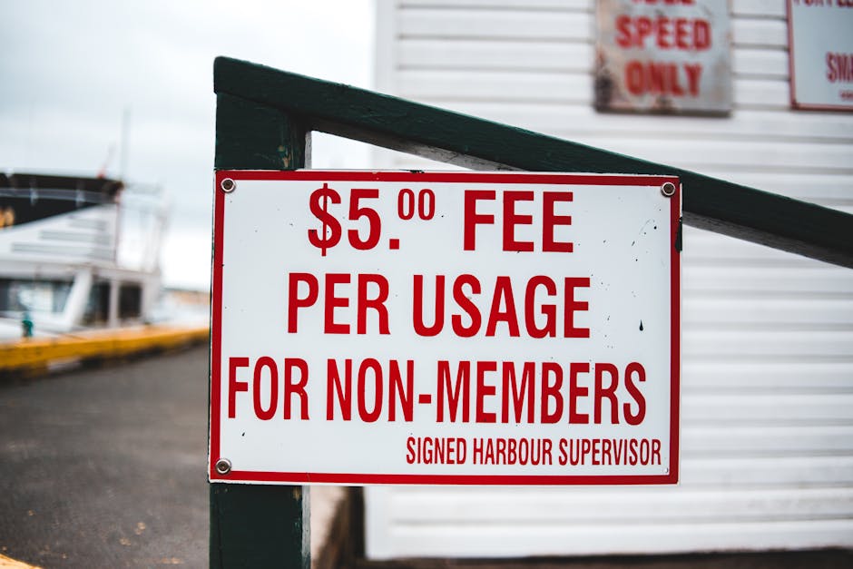 Close-up of a white and red sign attached to a dark green wooden post; the sign displays a fee of $5.00 per usage for non-members, with the text 'SIGNED HARBOUR SUPERVISOR' at the bottom. In the background, there are blurred images of a marina with boats docked along a quay, and a paved area leading to the water. The scene suggests a maritime or harbour environment, possibly relevant to house or item removals involving waterfront properties or boat transports. The photograph is taken during daytime under overcast skies, providing neutral lighting. This setting might be used in visual content related to relocation logistics near harbours or waterfronts, relevant to professional removals or moving services like those offered by Man with Van Blackheath.