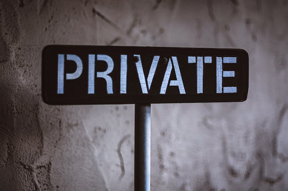 A close-up of a rectangular black metal sign with the word 'PRIVATE' displayed in illuminated blue capital letters, mounted on a vertical pole against a textured, off-white wall. The sign appears to be outdoors or in an entryway, indicating restricted access or privacy. In the context of house removals, such signage may be visible on a residential property during a home relocation, where furniture, boxes, and packing materials are being prepared for transport. The scene suggests a formal boundary or security measure, with the focus on the process of packing and moving, supported by the presence of equipment or environment typical in moving procedures. This image aligns with services provided by [COMPANY_NAME], such as careful handling of belongings and respect for property boundaries during the loading process.
