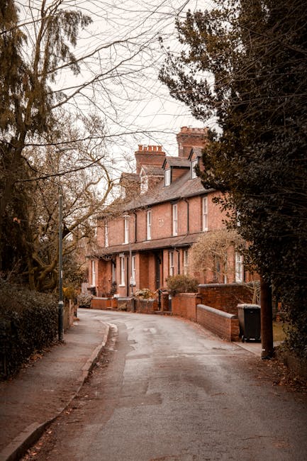 A residential street scene during daytime showing a narrow road lined with parked cars on both sides, including small hatchbacks, sedans, and delivery vans. The street is situated in a suburban area w