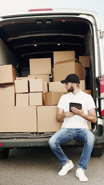 A middle-aged man wearing glasses, a light blue shirt, and dark trousers is seated on a wooden chair outside a white van used for house removals, taking a break during a packing and moving process. Th