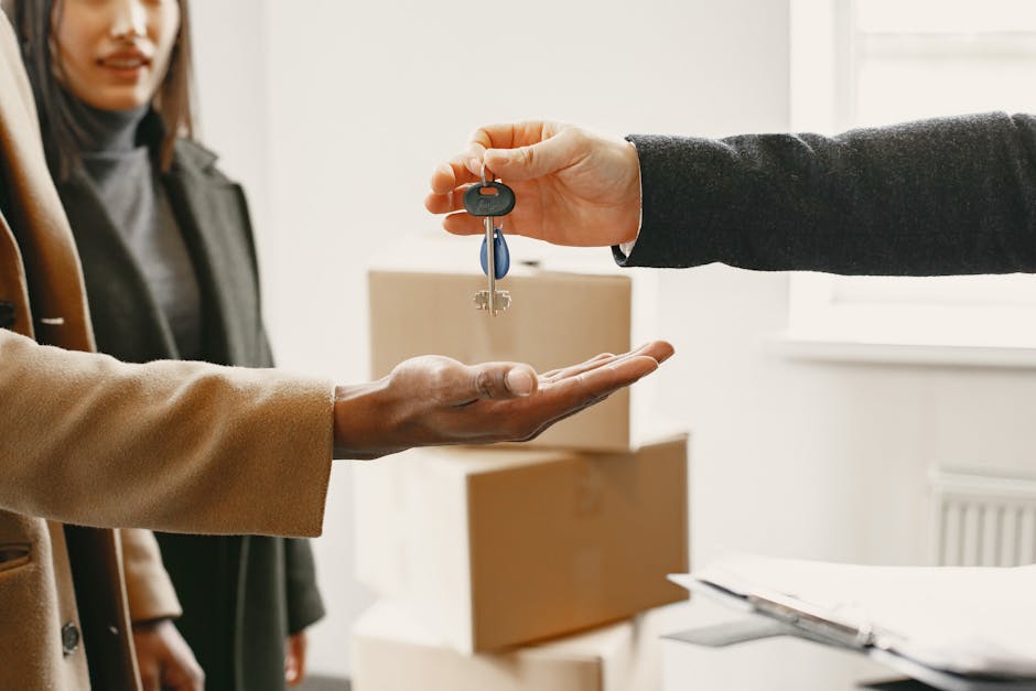 A person wearing a dark suit is handing over a set of keys to another individual dressed in a beige coat in an indoor setting, likely an office or property location. The person receiving the keys has their open palm outstretched, and behind them, a woman with shoulder-length dark hair is observing the exchange. In the background, there are cardboard boxes and packing materials on a table or countertop, indicating preparations for house relocation or furniture transport. The lighting is natural, coming from a nearby window, and the scene reflects the handover process involved in house removals or moving services, as provided by companies like Man with Van Blackheath. This moment illustrates the transfer of access or ownership, which is a common part of home moving and relocation logistics.