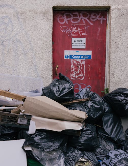 A stack of black rubbish bags filled with waste materials is piled against a red metal door with graffiti and signage, located on an exterior wall of a building. The waste includes cardboard boxes, plastic packaging, and miscellaneous refuse, some partially opened to reveal contents such as foam or furniture packaging remnants. The bags are situated on the pavement near the doorway, with some leaning against the door frame. The door, which may be part of a commercial or residential property, displays graffiti tags and warning signs, including one that indicates 'NO MONEY' and another that instructs to 'Keep clear.' The overall scene suggests a temporary accumulation of waste, potentially related to house or furniture removals or clearance activities, with visible effort to manage bulky waste disposal as part of house relocation or moving logistics. Man with Van Blackheath's services could involve packing, moving, or waste removal to facilitate property clearance and home relocation.