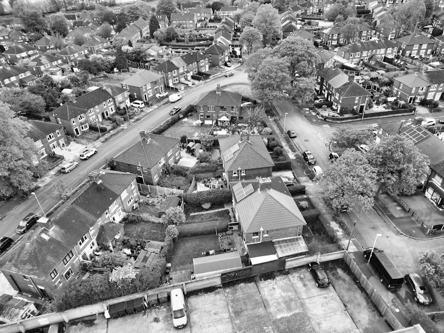An aerial black and white photograph of a residential neighbourhood showing multiple houses, gardens, and parked cars along the streets. In the foreground, there is a flat roof structure with visible windows and an adjacent yard with tools, a small shed, and landscaping. Behind, a narrow street curves through the area with several vehicles parked along the pavement, including a large van that is being loaded with household furniture and packing boxes wrapped in plastic and cardboard. The scene shows a typical urban setting for home relocation or furniture transport, with a focus on the loading process facilitated by [COMPANY_NAME], situated within the vicinity of houses surrounded by trees and greenery. The environment is well-lit with natural daylight, emphasizing the orderly street layout and the organization involved in professional removal services, such as packing, loading, and transportation within the area.