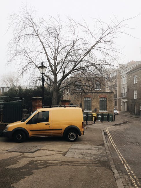 A yellow cargo van operated by Man with Van Blackheath is parked on a street near a residential building with grey brick walls and large windows. The van is positioned close to the curb, facing left, with no visible signage. In the background, a leafless tree stands behind a black lamp post, with its branches extending over the van. Adjacent to the van, there are several green wheelie bins lined up against the building. The street has a gentle slope, with double yellow lines on the right side, indicating parking restrictions. The scene appears to be part of a home relocation process, with the van likely used for furniture transport or packing and moving activities associated with house removals, supporting local move logistics. The environment is overcast, and the area looks quiet and residential, consistent with typical Blackheath street scenes.