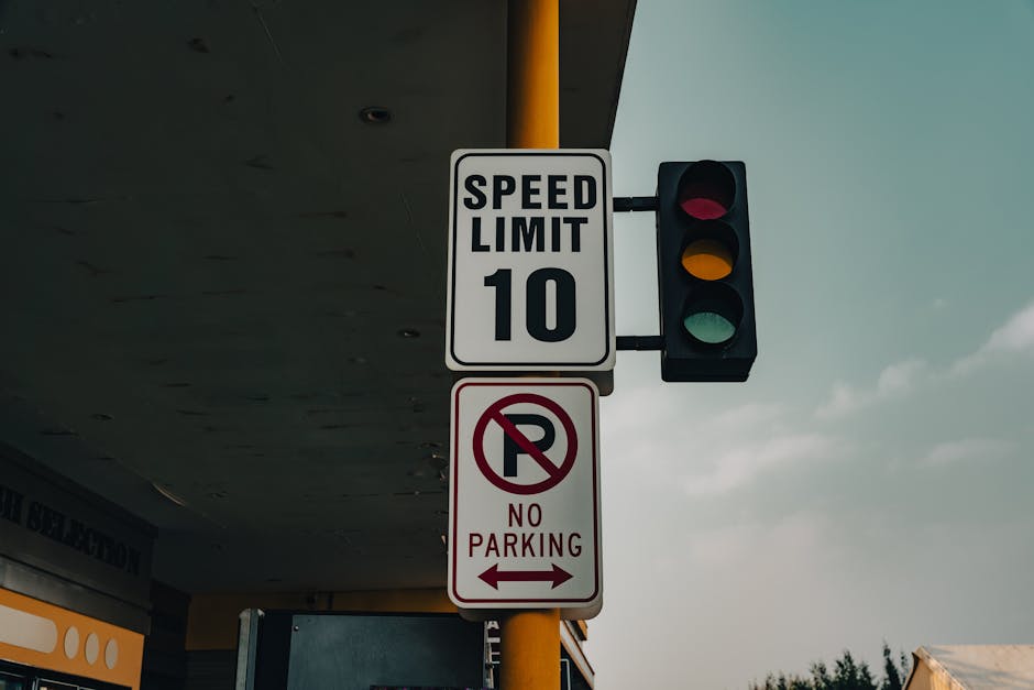 A traffic light with red, yellow, and green signals mounted on a black pole with a yellow accent featuring two rectangular street signs attached; the top sign displays a speed limit of 10 miles per hour in black text on a white background, while the lower sign, with a red circle and slash over a letter 'P', indicates no parking with arrows pointing left and right. The scene is outdoors under a grey, overhanging structure, possibly a petrol station canopy, with a partly cloudy sky in the background. The image reflects urban traffic regulation and parking restrictions relevant to house removals and transportation services offered by Man with Van Blackheath, situated in Lewisham SE3, and highlights the importance of adhering to parking and loading rules during furniture transport and home relocation processes.