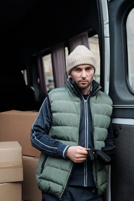 A man with light skin, wearing a beige knit beanie, a navy blue jacket with white piping, and a light green padded vest, stands inside the open rear door of a moving van. He holds a black strap or sec