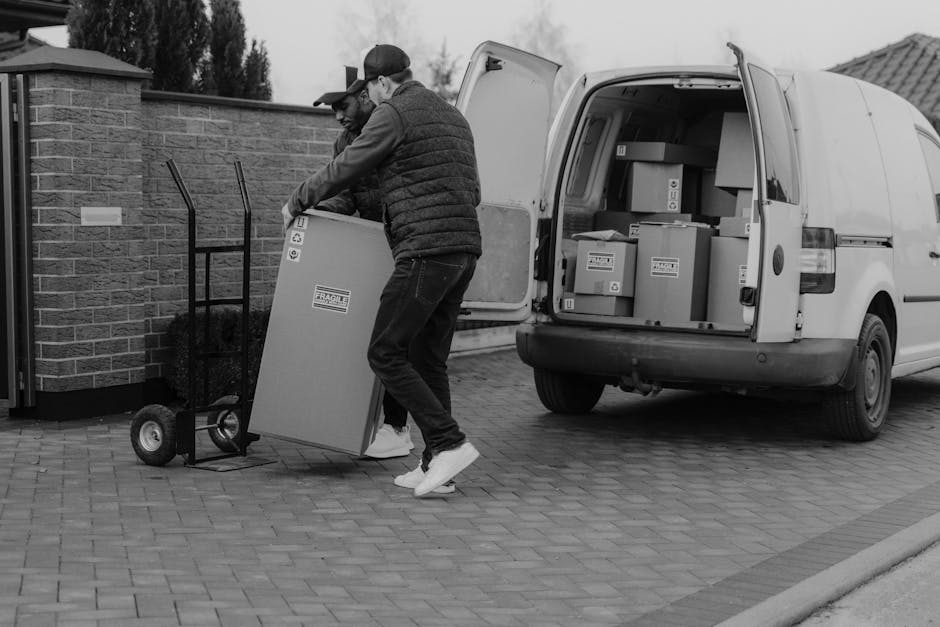 A man with light skin, wearing a beige knit beanie, a navy blue jacket with white piping, and a light green padded vest, stands inside the open rear door of a moving van. He holds a black strap or sec
