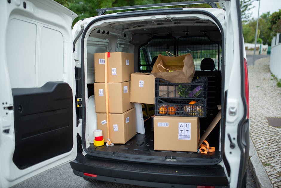 The rear cargo area of a white moving van, likely used for home relocation, is shown with the back door open. Inside, there are several brown cardboard boxes, some stacked vertically and others placed side by side, each with white labels and barcodes, indicating they are part of packing and moving preparations by Man with Van Blackheath. A black plastic crate filled with fresh produce, including oranges and purple vegetables, is positioned on the right side, with a brown paper bag on top. Additionally, a roll of plastic wrapping and packing materials are visible. The van is parked on a paved street with a curb on the right side, and the environment is outdoors with trees and residential buildings in the background. This image illustrates the loading process typical of furniture transport and packing for house removals, showcasing the careful organization of packed items inside a vehicle prepared for a local household move.