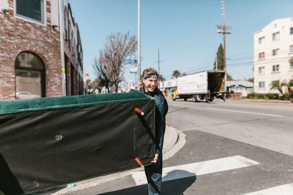 A man with long blonde hair, wearing a patterned headband and dark clothing, is outdoors on a street corner, smiling while carrying a large, padded, black and green furniture moving blanket. The blanket is secured with orange and black straps, and he holds one side of it as he assists with a home relocation. In the background, there is a white delivery truck parked partially on the road, a brick building with arched windows to the left, and a white multi-story building further down the street. The street has a pedestrian crossing and utility poles with power lines, with a clear blue sky overhead. The scene depicts the loading process involved in furniture transport and house removals, with [COMPANY_NAME] providing professional packing and moving services relevant to house moves in Blackheath, SE3.