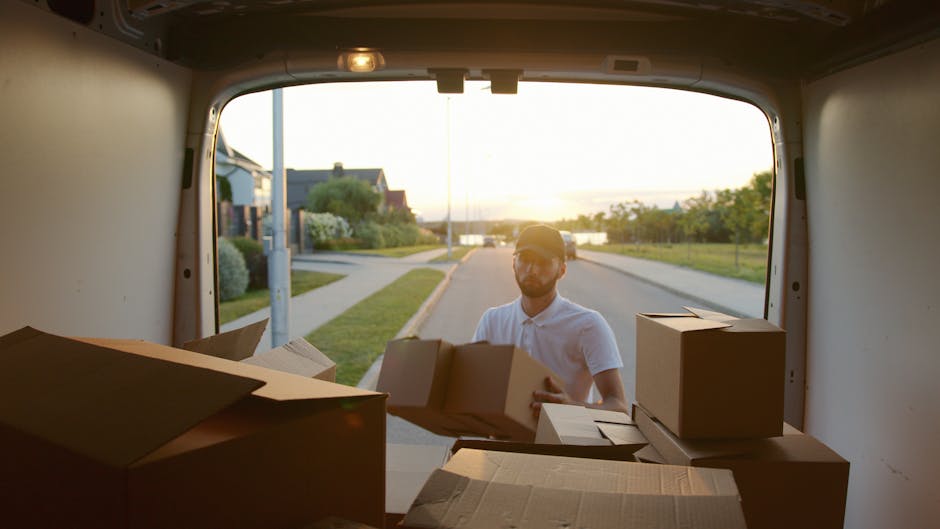 The rear cargo area of a white moving van, likely used for home relocation, is shown with the back door open. Inside, there are several brown cardboard boxes, some stacked vertically and others placed side by side, each with white labels and barcodes, indicating they are part of packing and moving preparations by Man with Van Blackheath. A black plastic crate filled with fresh produce, including oranges and purple vegetables, is positioned on the right side, with a brown paper bag on top. Additionally, a roll of plastic wrapping and packing materials are visible. The van is parked on a paved street with a curb on the right side, and the environment is outdoors with trees and residential buildings in the background. This image illustrates the loading process typical of furniture transport and packing for house removals, showcasing the careful organization of packed items inside a vehicle prepared for a local household move.