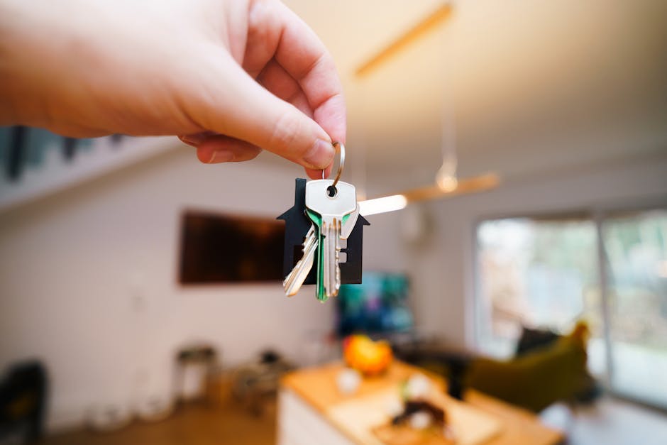 A close-up of a person's hand holding a set of house keys attached to a keyring, with several metal keys and a black plastic key fob. The background shows a modern, well-lit living room with a flat-screen television mounted on the wall, a wooden dining table with an orange fruit bowl, and a sofa near large windows that let in natural light. The scene depicts the initial stage of home relocation or moving process, highlighting key handover within a residential property, consistent with house removal services provided by Man with Van Blackheath, specialists in packing, loading, and furniture transport.