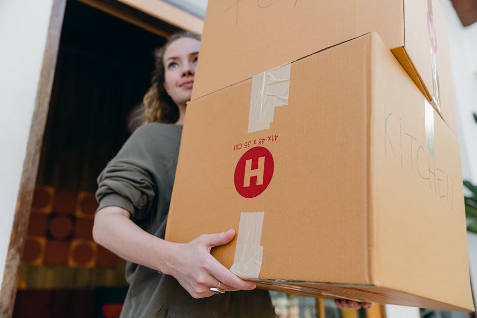 A woman with light skin and wavy brown hair, dressed in a gray long-sleeve top, is inside a property holding a cardboard box with both hands, preparing to load or carry it. The box features packing tape and a printed red 'H' symbol, indicating it contains fragile items. Behind her, part of a wooden door frame and a wall with a coat rack or storage area with cylinders of material are visible. The environment suggests an indoor setting during a house relocation, with natural lighting illuminating her face and the box. The scene illustrates the process of packing and moving, with the woman engaged in handling household items as part of a professional removals service provided by Man with Van Blackheath, who specialise in home removals and furniture transport for owners and landlords, especially during night and weekend moves around Blackheath.