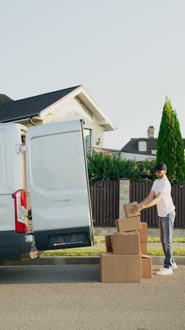 A person holds a remote control drone in their hands, guiding it to capture footage of a house removal process. In the drone's live camera view displayed on a smartphone mounted on top of the remote, 