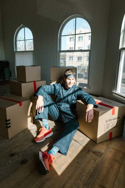 A young man sitting on a wooden floor inside a bright room with large arched windows, surrounded by several sealed cardboard boxes with red packing tape, indicating a home relocation or packing process. The man is wearing casual clothing, including a dark blue jacket, dark blue trousers, bright orange sneakers, and blue socks with striped patterns. Sunlight streams through the windows, casting shadows across the floor, and the man is smiling while leaning back against one of the boxes, suggesting a moment during furniture transport or packing and moving activities. This scene reflects the logistics involved in house removals, supported by the presence of packing materials and the natural lighting typical of the interior environment managed by a professional removals service like Man with Van Blackheath.
