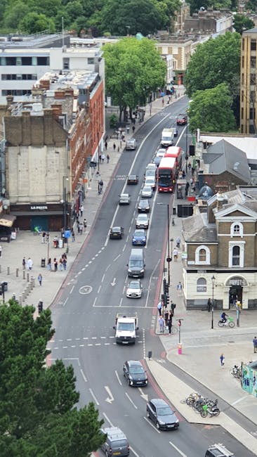 An aerial view of a busy urban street with parked and moving cars, including a red double-decker bus, navigating through a cityscape lined with a mix of brick and modern buildings. Pedestrians are walking along the sidewalks, some crossing at designated crossings, while others stand near storefronts. Several bicycles are parked on the pavement near a small, classic-style building with a gabled roof. Tall, leafy green trees are visible along the street, providing shade and greenery in the city environment. In the distance, additional buildings and trees form the backdrop, with the street curving out of view. The scene captures typical city movement and activity, relevant to home relocation and furniture transport contexts, as part of the comprehensive moving services offered by Man with Van Blackheath.