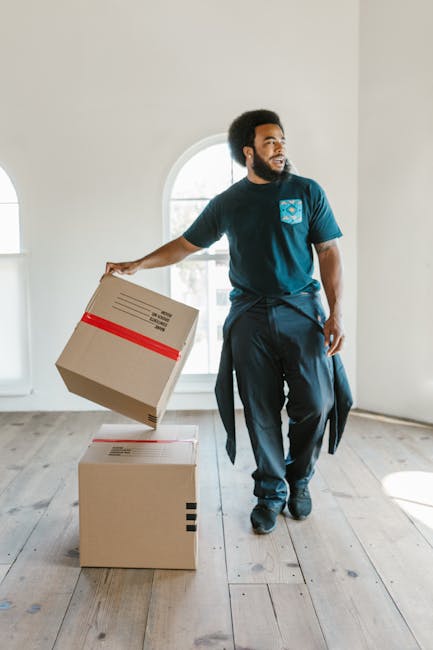 A young man sitting on a wooden floor inside a bright room with large arched windows, surrounded by several sealed cardboard boxes with red packing tape, indicating a home relocation or packing process. The man is wearing casual clothing, including a dark blue jacket, dark blue trousers, bright orange sneakers, and blue socks with striped patterns. Sunlight streams through the windows, casting shadows across the floor, and the man is smiling while leaning back against one of the boxes, suggesting a moment during furniture transport or packing and moving activities. This scene reflects the logistics involved in house removals, supported by the presence of packing materials and the natural lighting typical of the interior environment managed by a professional removals service like Man with Van Blackheath.